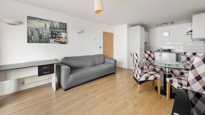 Un espace de vie moderne avec un canapé gris, une table à manger en verre et des chaises à motifs à carreaux, un coin cuisine et une œuvre d’art représentant un paysage urbain accrochée au mur.