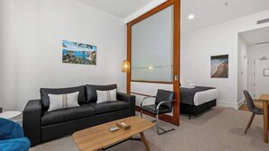 Une chambre d’hôtel moderne avec un canapé noir, une table basse en bois, un lit recouvert d’une couette blanche et un grand miroir.