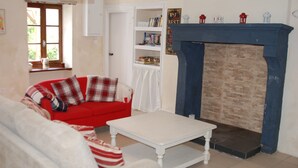 Een woonkamer met een rode bank, een witte fauteuil en een witte salontafel. Er is een open haard met een blauwe schouw en een boekenplank vol boeken en decoratieve objecten.