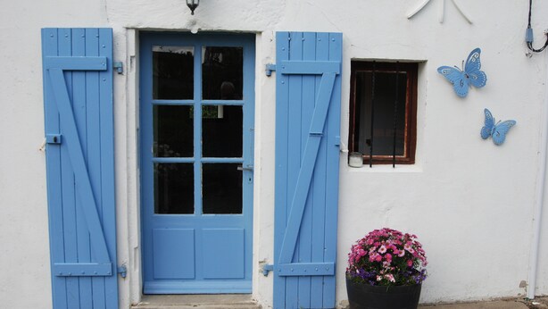 Een wit gebouw met een blauwe deur en luiken, een klein raam en een bloempot met roze bloemen.