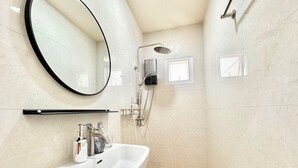 Un baño moderno con un lavamanos blanco, un espejo redondo, un cabezal de ducha de mano y una ventana pequeña.