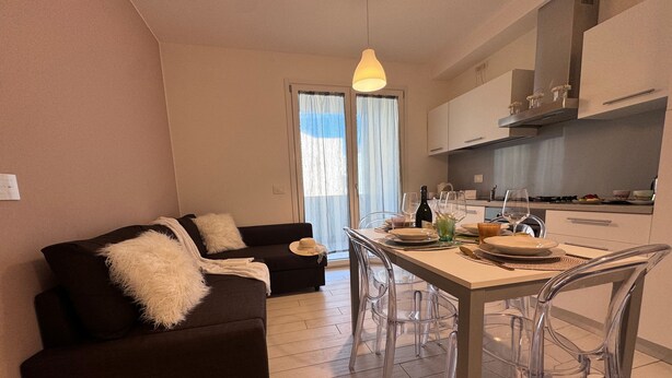 A modern living room with a dark brown sofa, a dining table set with plates and glasses, and a kitchen area with cabinets and a hanging light fixture. - Casa Helios by Ca-sa Check (Chioggia)