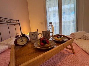 Una bandeja de madera con alimentos para el desayuno, dos tazas con texto, un reloj y una botella sobre una cama con vistas al exterior.