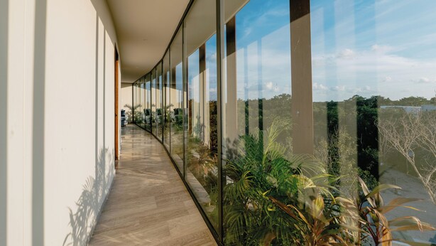 Un pasillo con paredes de vidrio y suelo de madera, que conduce a vistas de vegetación y un cielo despejado.