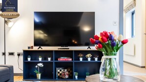 Una sala de estar moderna con un televisor de pantalla plana grande, un mueble azul para entretenimiento con plantas y juegos de mesa, un sofá gris y una pequeña mesa redonda con un jarrón de tulipanes.