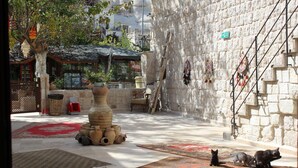 Un patio con muros de piedra, suelo de baldosas y un gran jarrón con plantas. Hay alfombras en el suelo y una pequeña mesa con sillas.