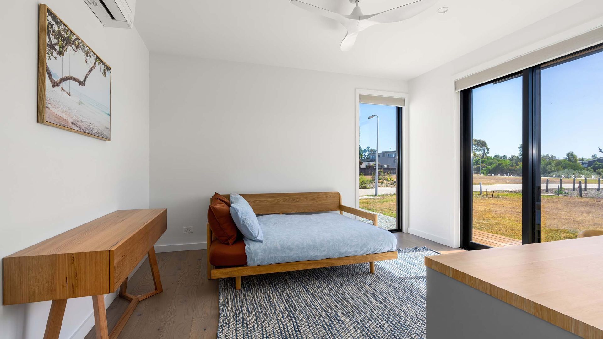 A modern bedroom with a bed, a wooden desk, and a large window offering a view of the outdoors.