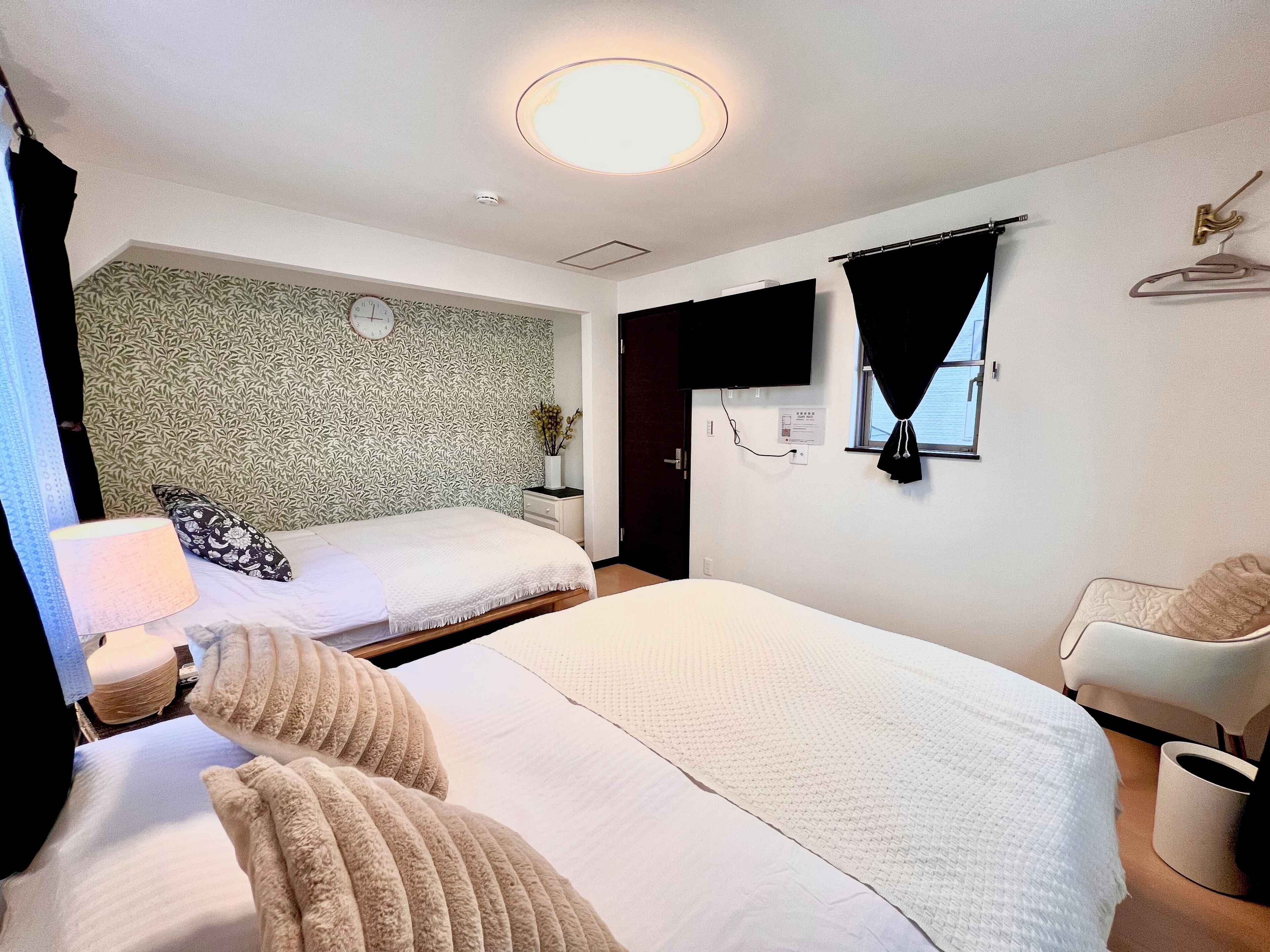 A hotel room with two beds, a bedside table, a TV, a window with curtains, and a wall clock.