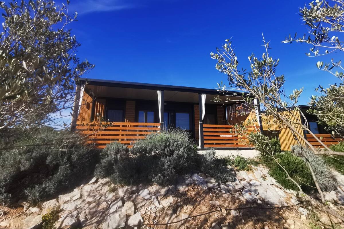 Una cabaña moderna con una terraza de madera, rodeada de vegetación natural y un cielo azul despejado.