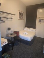 Habitación de hotel con cama, una mesita con lámpara, una silla, perchero y una puerta con letrero.