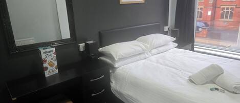 A hotel room with a bed, a bedside table with a mirror, a small table with a box of food, and a window with a view of buildings.
