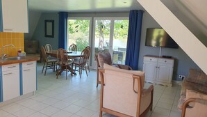 A dining area with a table and chairs, a kitchen with a sink and cabinets, a television on a stand, and a view of greenery through a glass door. - Rural 2 bedroom fully equiped Chalet (1 of 2) by lake near Chaillac sleeps max 6 (Chaillac)