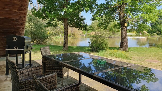 An outdoor patio with a glass table and wicker chairs, overlooking a pond and trees. - Rural 2 bedroom fully equiped Chalet (1 of 2) by lake near Chaillac sleeps max 6 (Chaillac)
