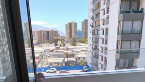A balcony with a cushioned chair and a small table, overlooking a cityscape with buildings and parked cars.