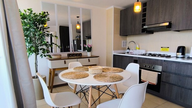 A modern kitchen with dark wood cabinets, a white countertop, and a round dining table with four white chairs. There are two pendant lights hanging above the table.