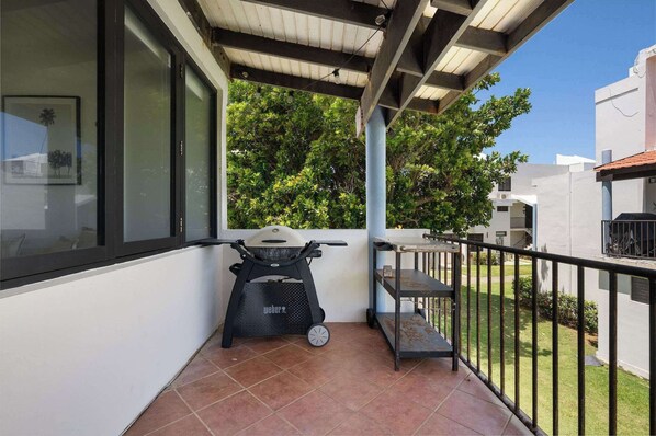 Un balcón con una parrilla Weber, un estante de metal y vistas a un jardín verde y edificios vecinos.