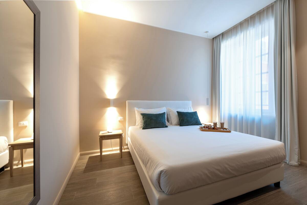Ein Hotelzimmer mit einem großen Bett, Nachttischen, einem Spiegel und einem Fenster mit Vorhängen.
