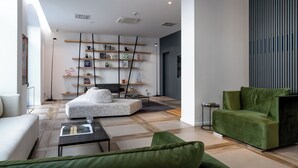 Een moderne woonkamer met een hoekbank, een groene fauteuil, een witte bank en een salontafel. Er staat een boekenplank met diverse decoratieve objecten en een vloerlamp.
