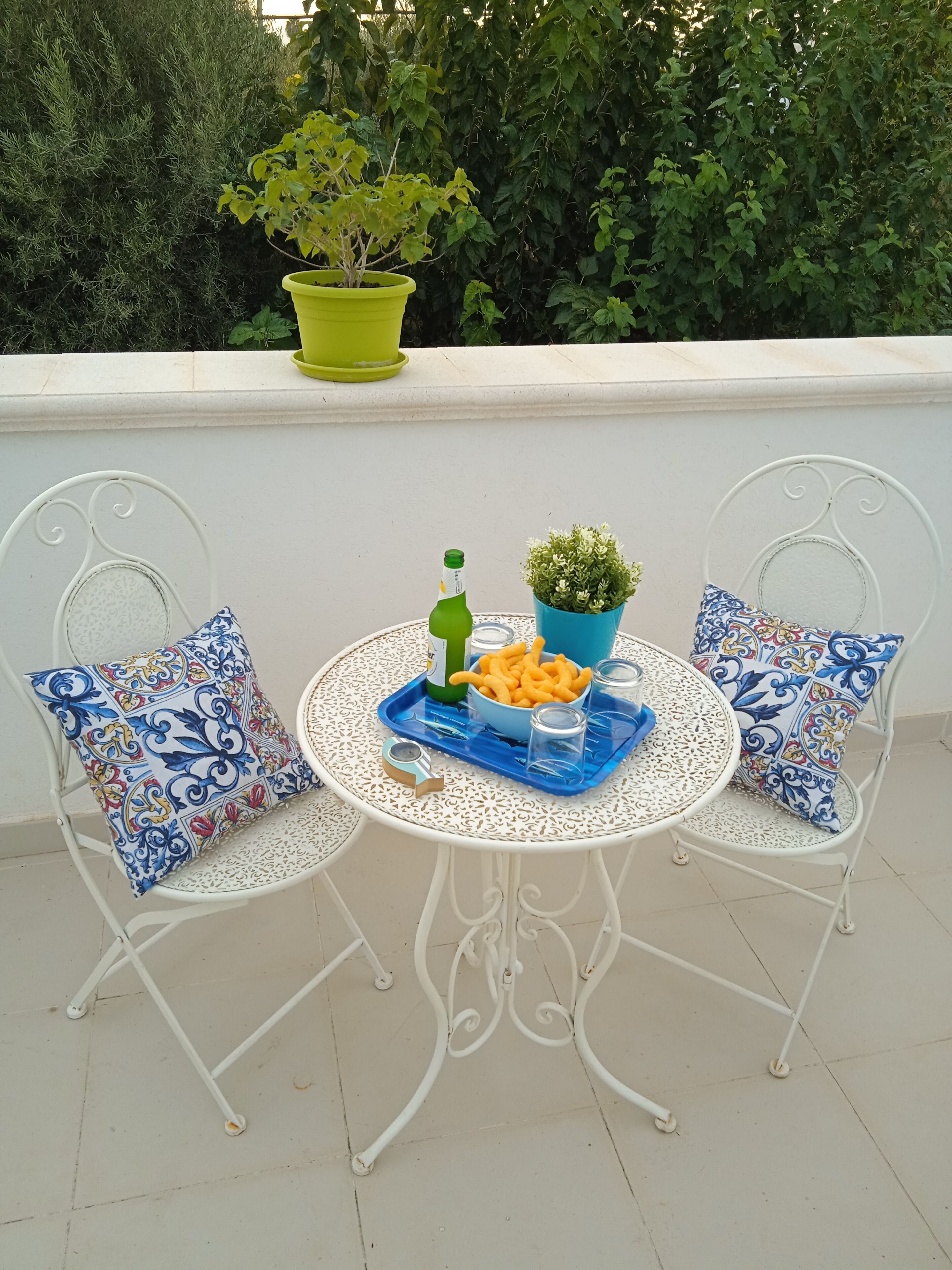 Una pequeña mesa exterior con dos sillas, una botella verde, un bol de aperitivos y una planta en maceta.