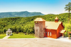 Uma cabana rústica com uma torre de pedra, rodeada por vegetação exuberante e montanhas.