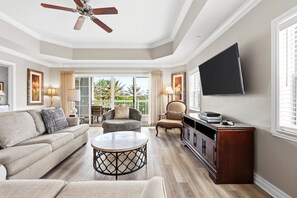 A spacious living room with a ceiling fan, a flat-screen TV mounted on the wall, a sofa, a coffee table, and a cabinet.