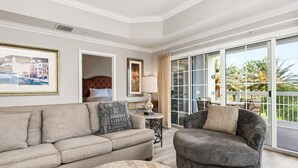 Un salon comprenant un canapé, un fauteuil et une table ronde. Une porte donne sur un balcon d’où l’on peut admirer des palmiers.