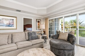 A living room with a sofa, armchair, and a round table. There is a door leading to a balcony with a view of palm trees.