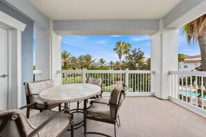 Une terrasse couverte avec une table ronde et des chaises, des rampes blanches et une vue sur une piscine et des palmiers.