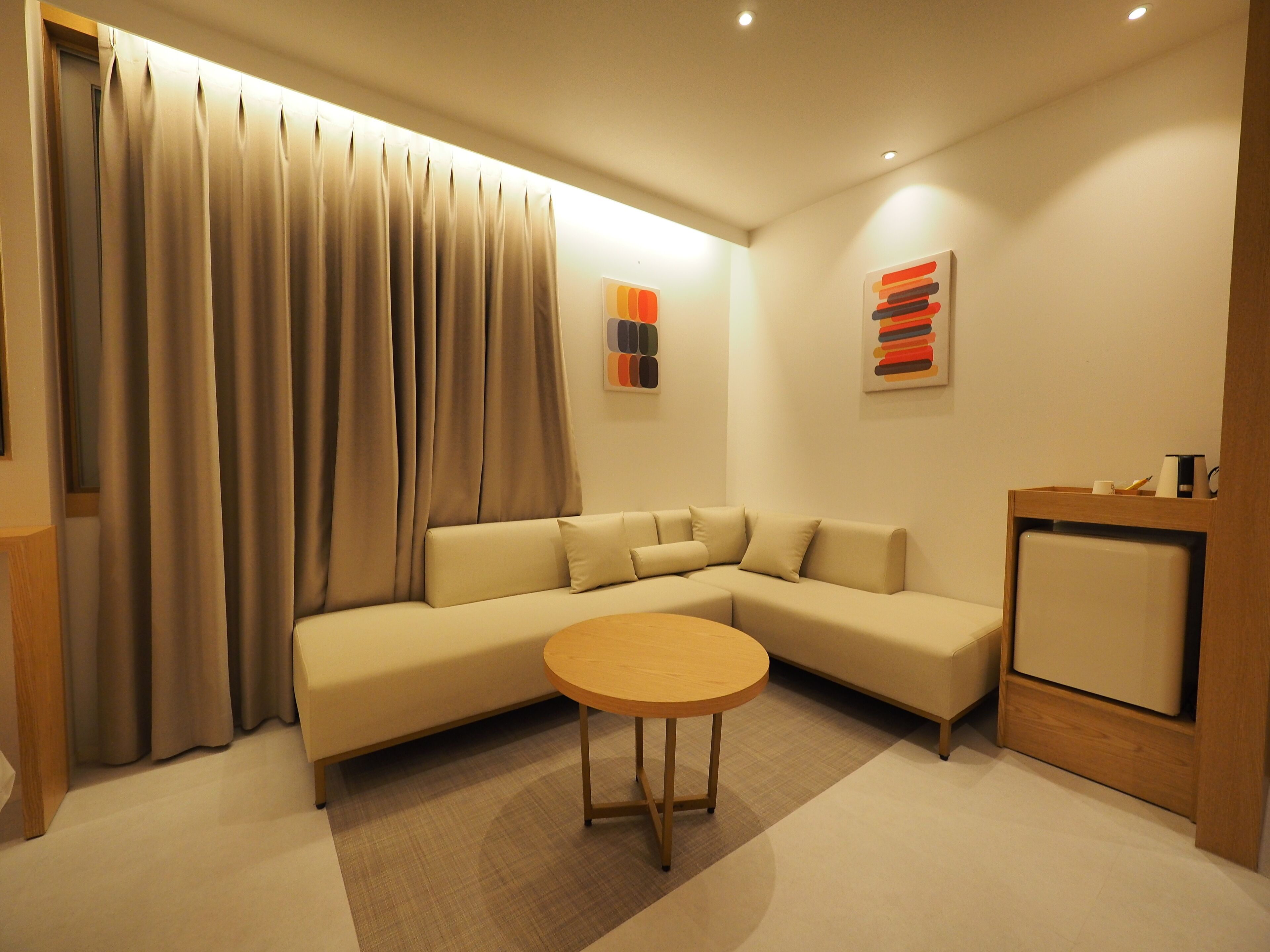 Una sala de estar moderna con un sofá seccional color beige, una mesa de centro redonda de madera y una mesita auxiliar de madera.