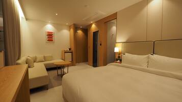 A hotel room with a large bed, a sofa, a small table, and a desk. There is a door and a wall-mounted light fixture.