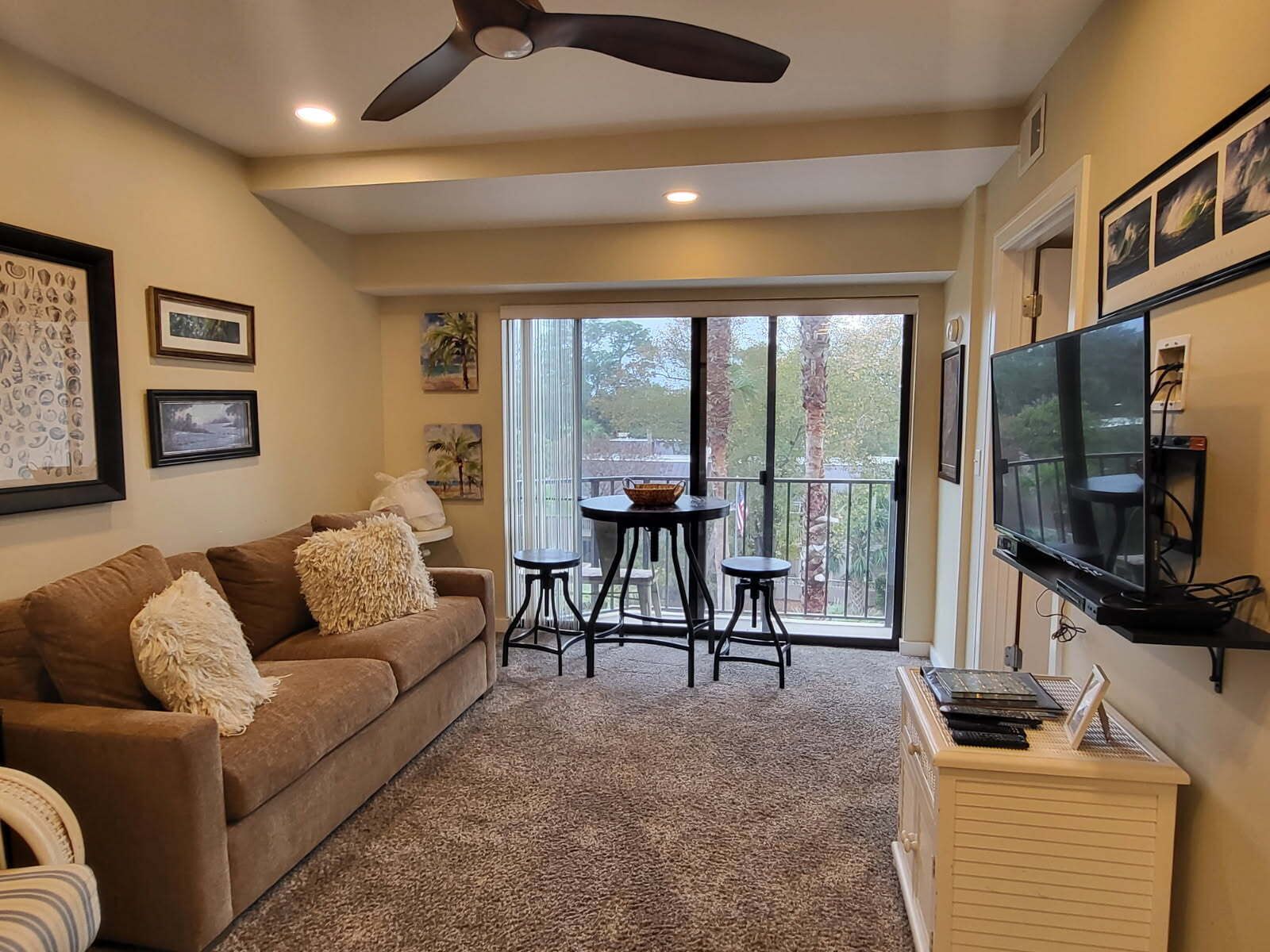 A living room with a brown sofa, a ceiling fan, a TV, and a balcony with a table and chairs.
