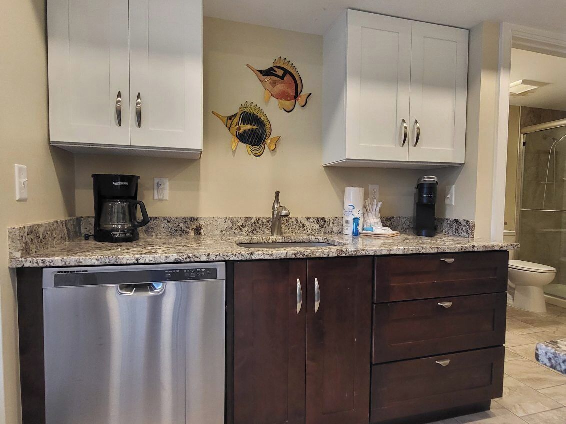 A kitchen with white upper cabinets, a dishwasher, dark lower cabinets, a granite countertop, a coffee maker, and a fish wall art piece.