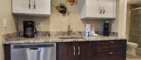 A kitchen with white upper cabinets, a dishwasher, dark lower cabinets, a granite countertop, a coffee maker, and a fish wall art piece.
