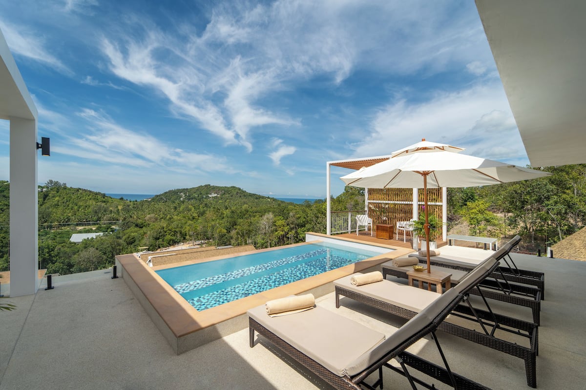 A rooftop infinity pool with lounge chairs, a pergola with an umbrella, and a view of the surrounding greenery and hills.