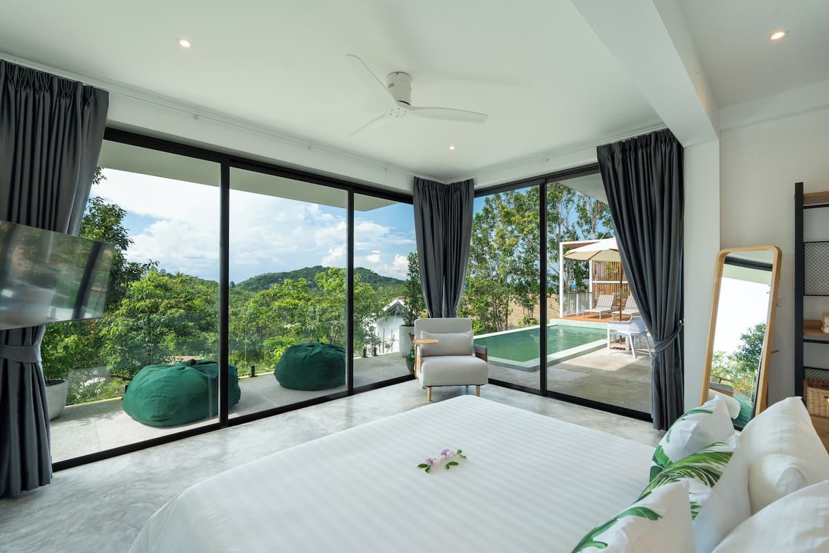 A modern hotel room with a large bed, a ceiling fan, and a view of a swimming pool and greenery.
