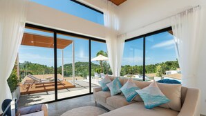 Una sala de estar moderna con un ventanal grande, un sofá con cojines decorativos, una mesa de madera y vistas a una piscina y una zona de estar exterior.