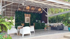 Un espacio al aire libre con sillas blancas, un sofá gris y una mesa de madera. Hay lámparas de mimbre colgantes y una pared cubierta de follaje verde.