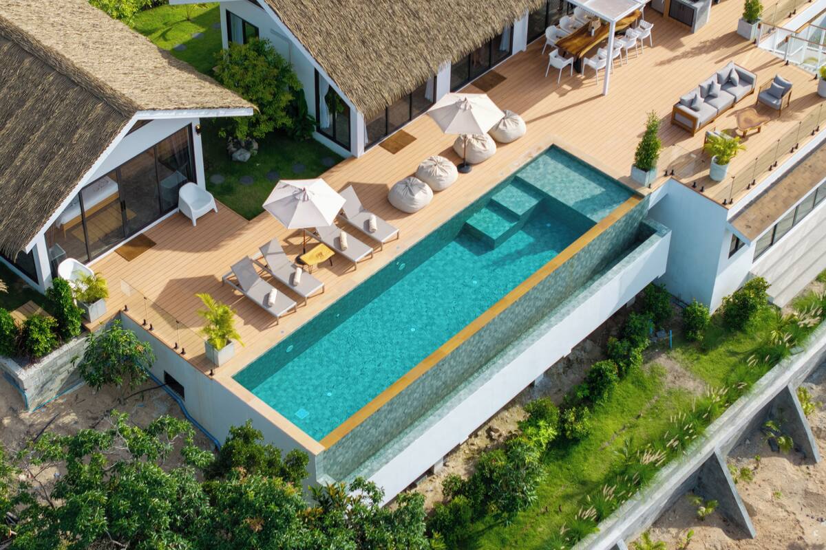 An aerial view of a modern resort with a large swimming pool, lounge areas, and thatched-roof buildings.