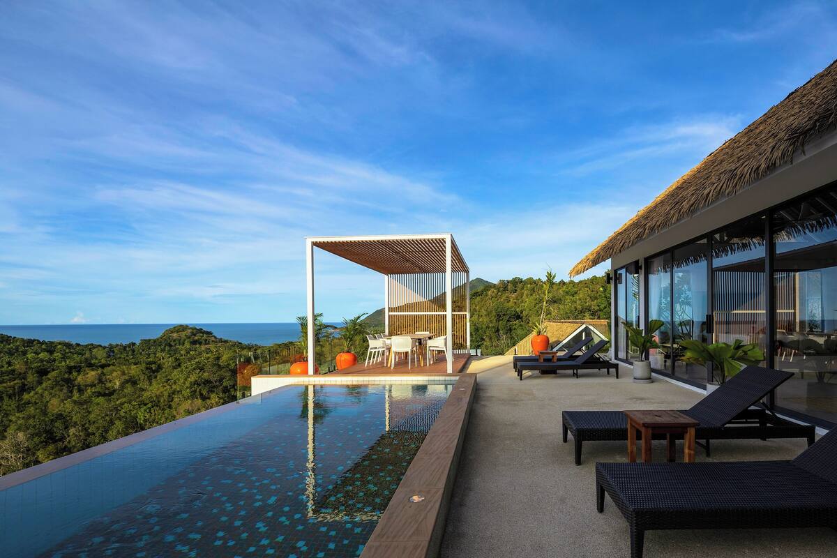 A modern outdoor pool area with lounge chairs, a wooden pergola, and a glass-enclosed structure overlooking a lush landscape.