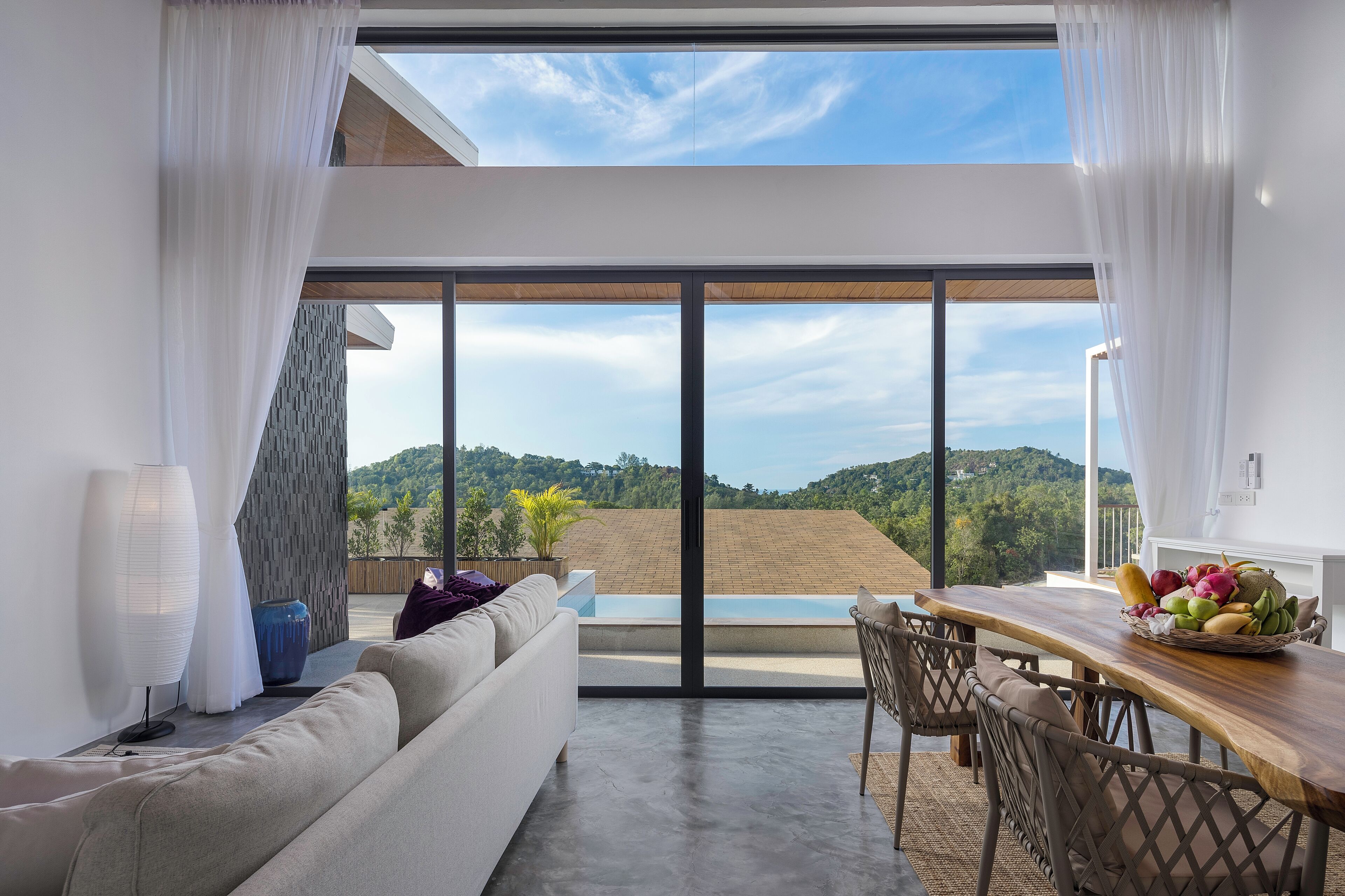 A modern living room with a large sliding glass door leading to a terrace, a wooden dining table with fruit, and a sofa with a potted plant.