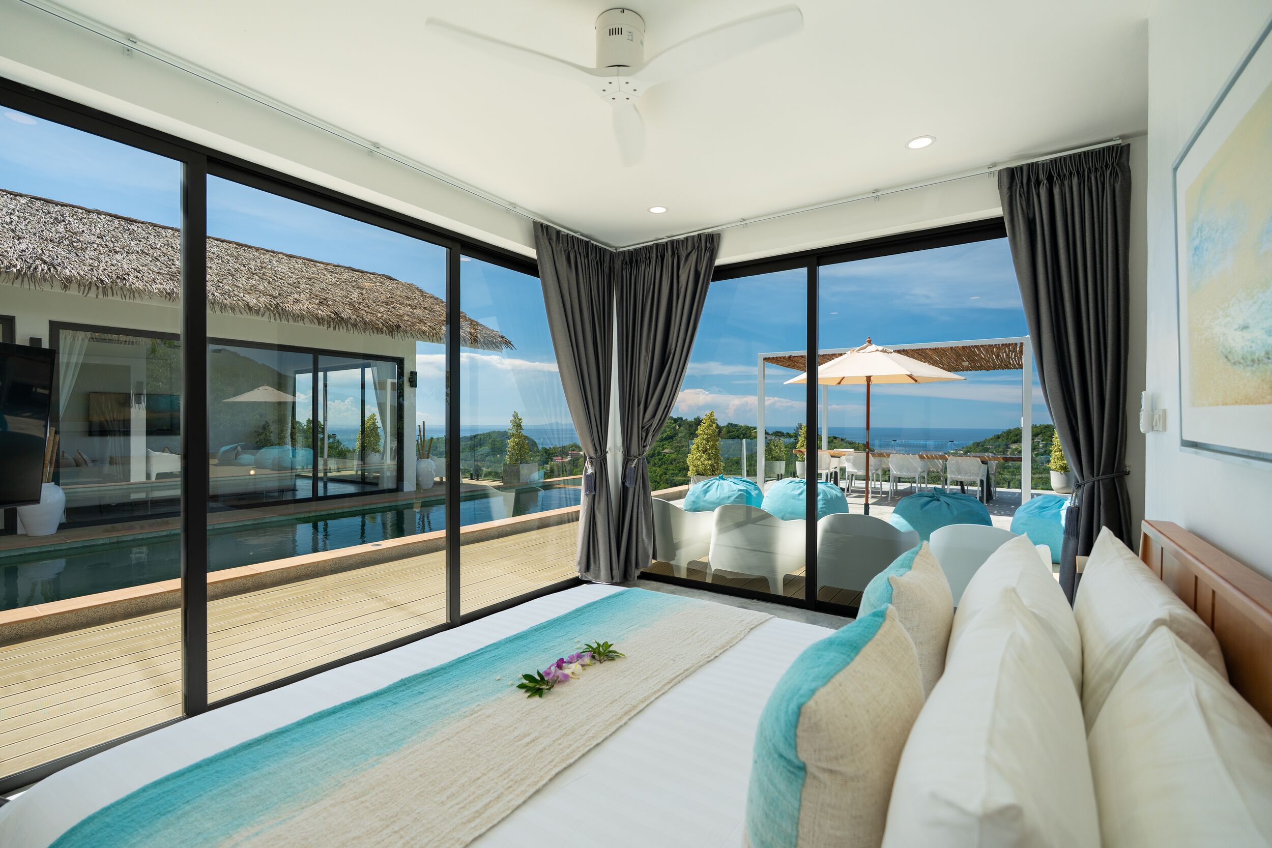 Camera da letto moderna con un letto grande, vista su piscina e area salotto esterna, e una ventola a soffitto.