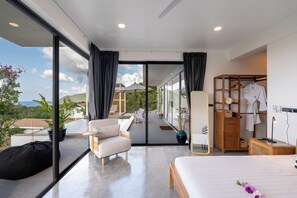 Una habitación de hotel moderna con una cama grande, una zona de estar con un sofá y una silla, un armario de madera y vistas al exterior.
