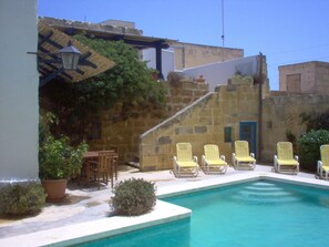 Egy medencetér, ahol napozóágyak és egy kis asztal található, melyet kőfalak és virágtartókban elhelyezett növények vesznek körül.