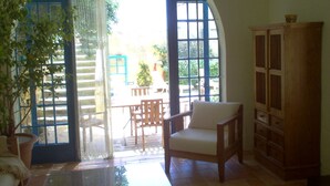 Un intérieur comprenant une chaise en bois, un meuble et, vue à travers une porte, un espace extérieur avec une table et des chaises.