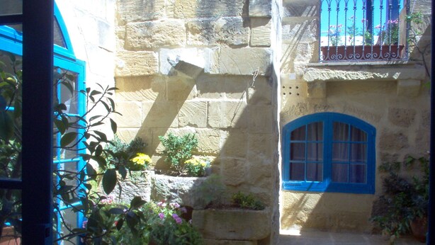 Une cour bordée de murs en pierre, avec une fenêtre bleue et des plantes en pot.