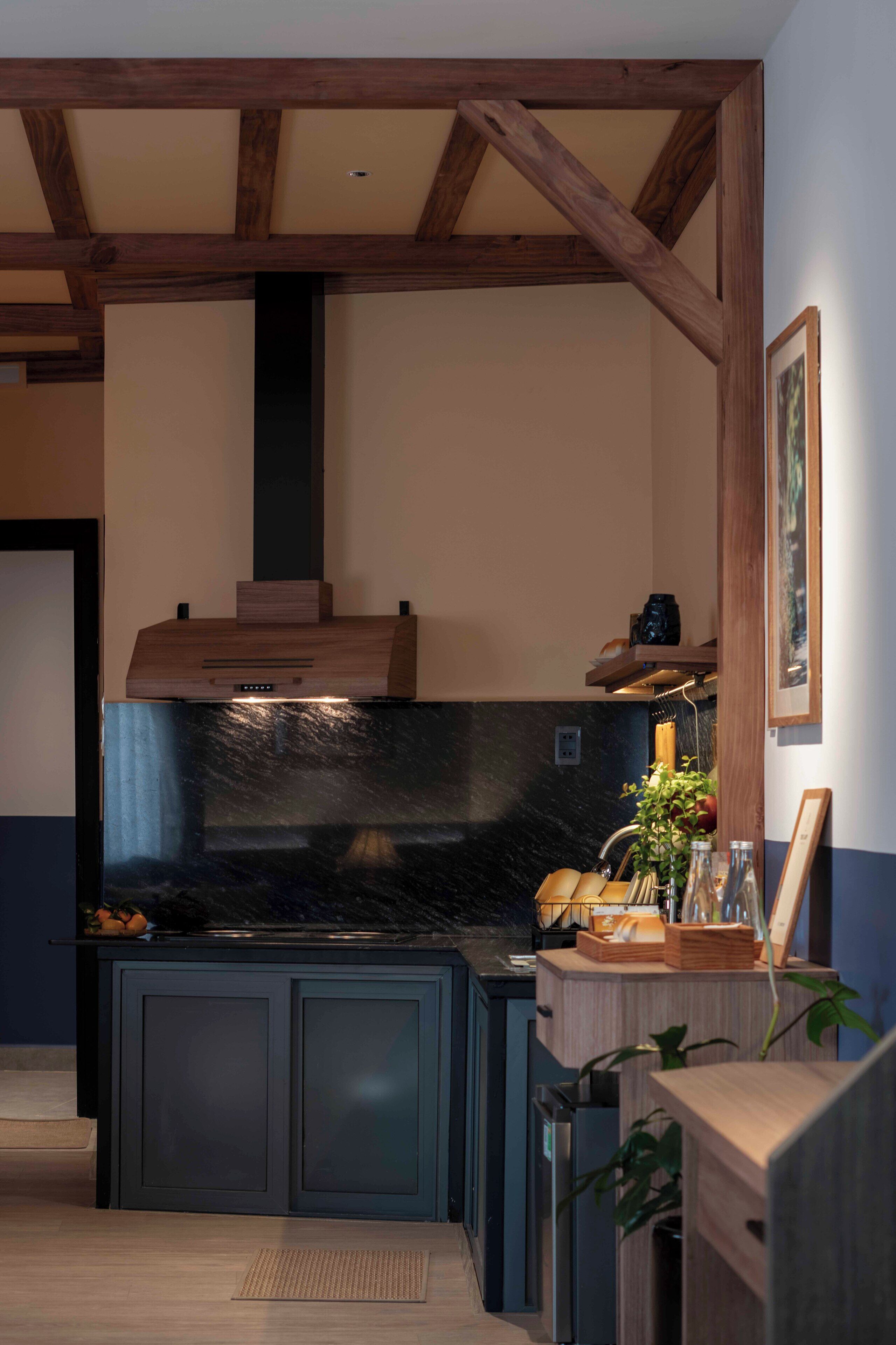 Una cocina con armarios oscuros, encimera negra, vigas de madera y una lámpara colgante.