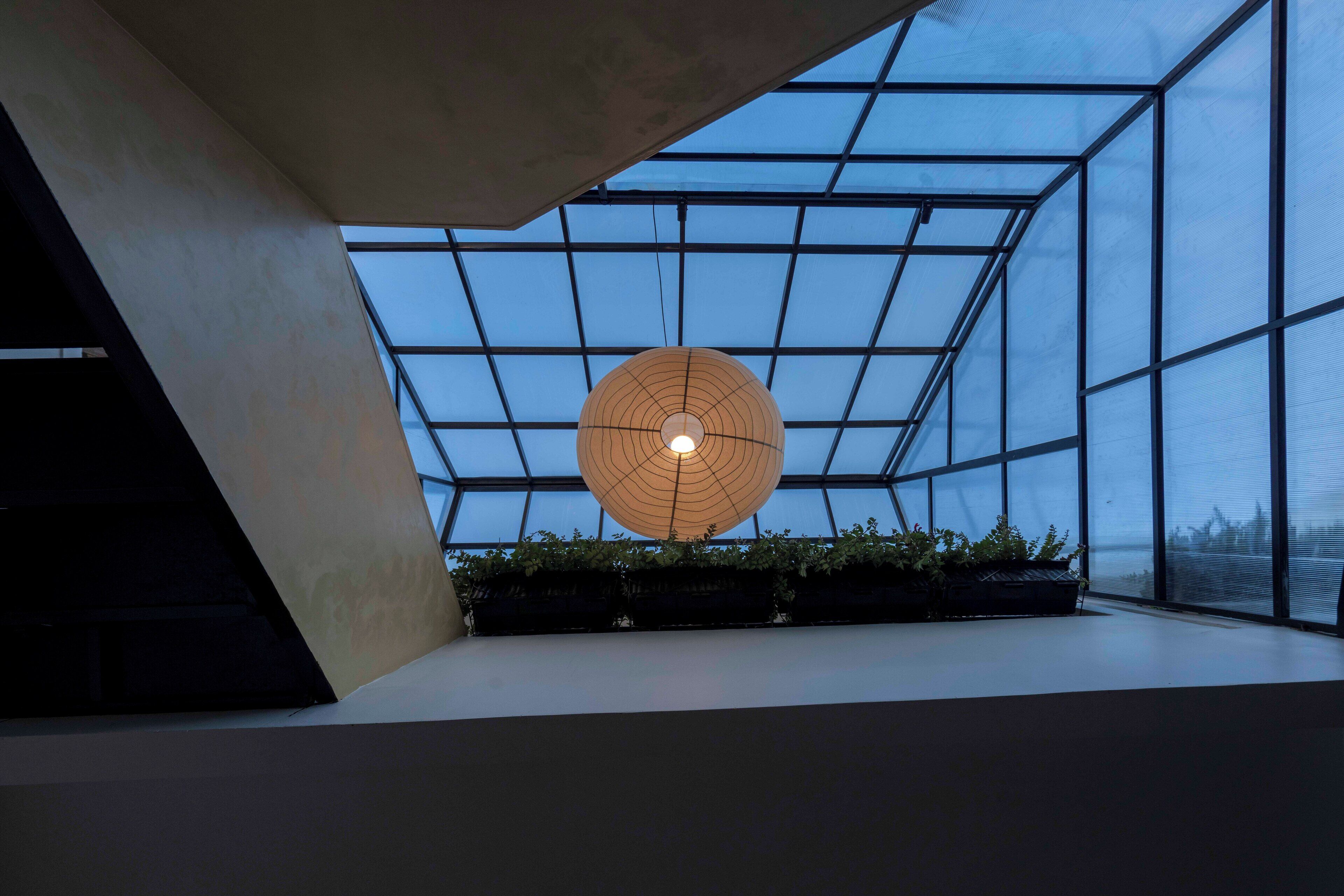 A modern interior with a geometric glass ceiling, a central hanging light fixture, and a row of planters along the glass wall.