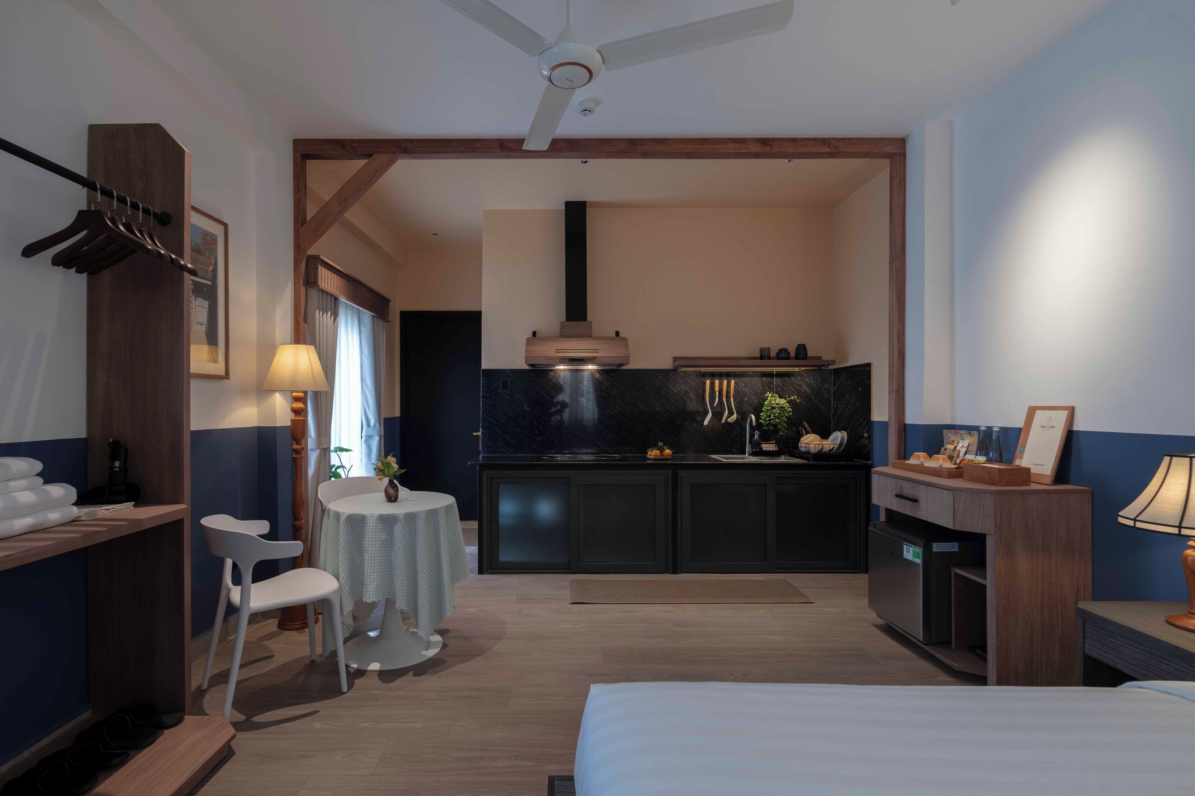 A modern hotel room with a bed, a desk, a chair, a small table, a kitchenette, and a ceiling fan.