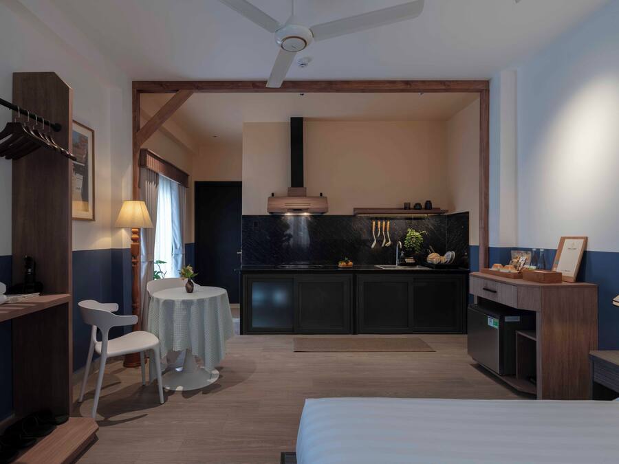 A modern hotel room with a bed, a desk, a chair, a small table, a kitchenette, and a ceiling fan.