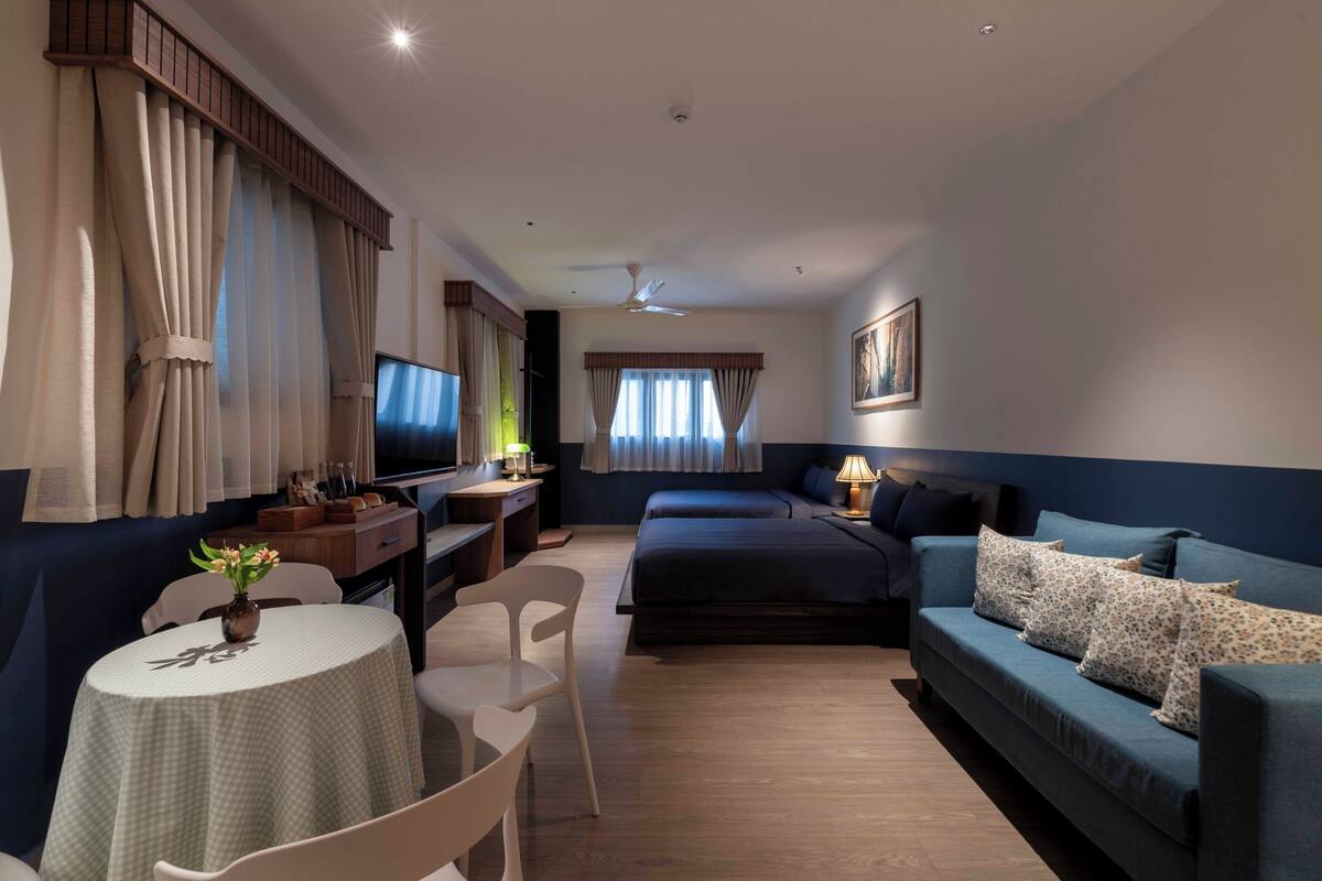 Une chambre d’hôtel comprenant un lit, un canapé, un bureau avec un ordinateur, une chaise et une petite table sur laquelle est posé un vase.